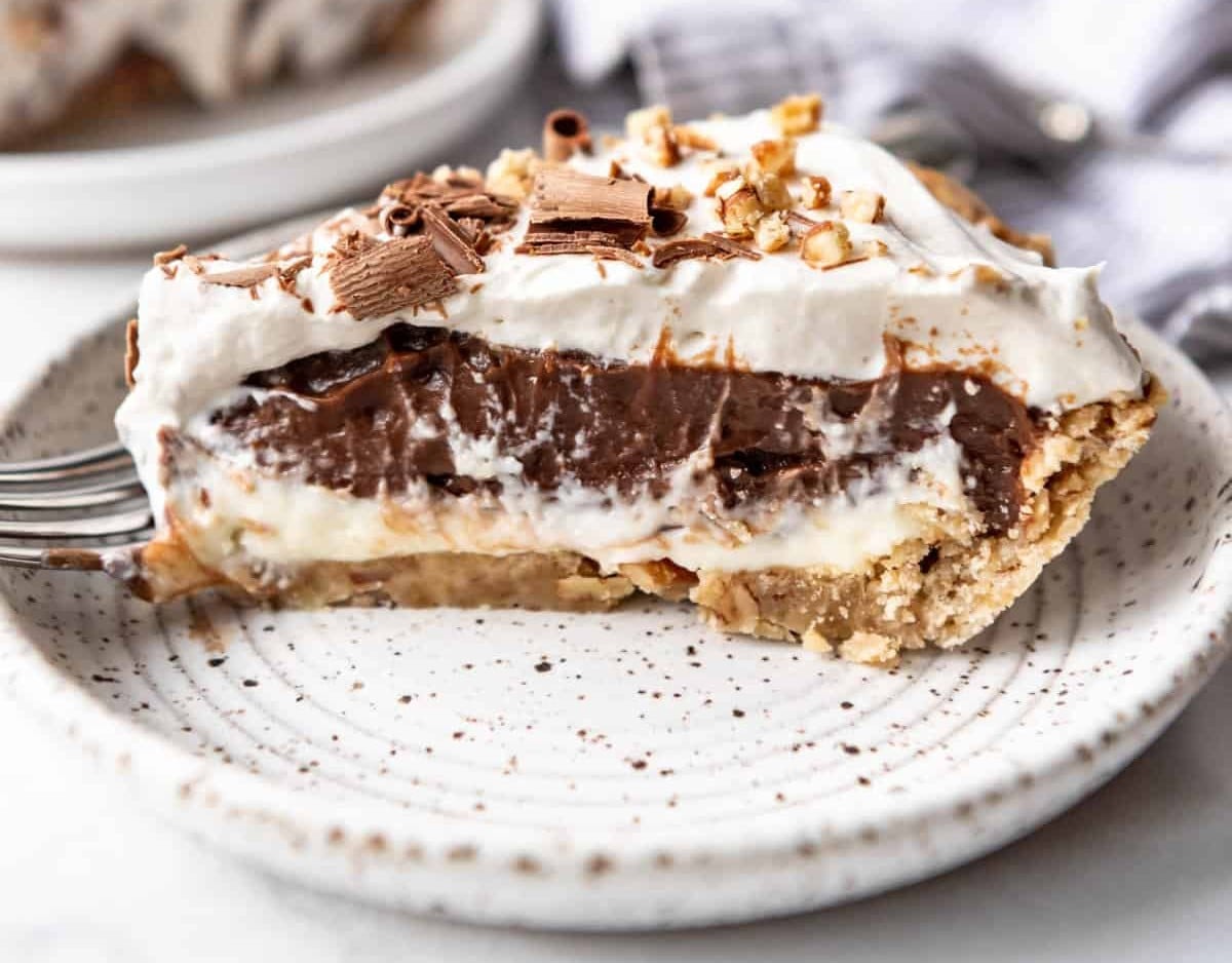 A slice of layered chocolate cream pie topped with whipped cream, chocolate shavings, and chopped nuts on a speckled plate.