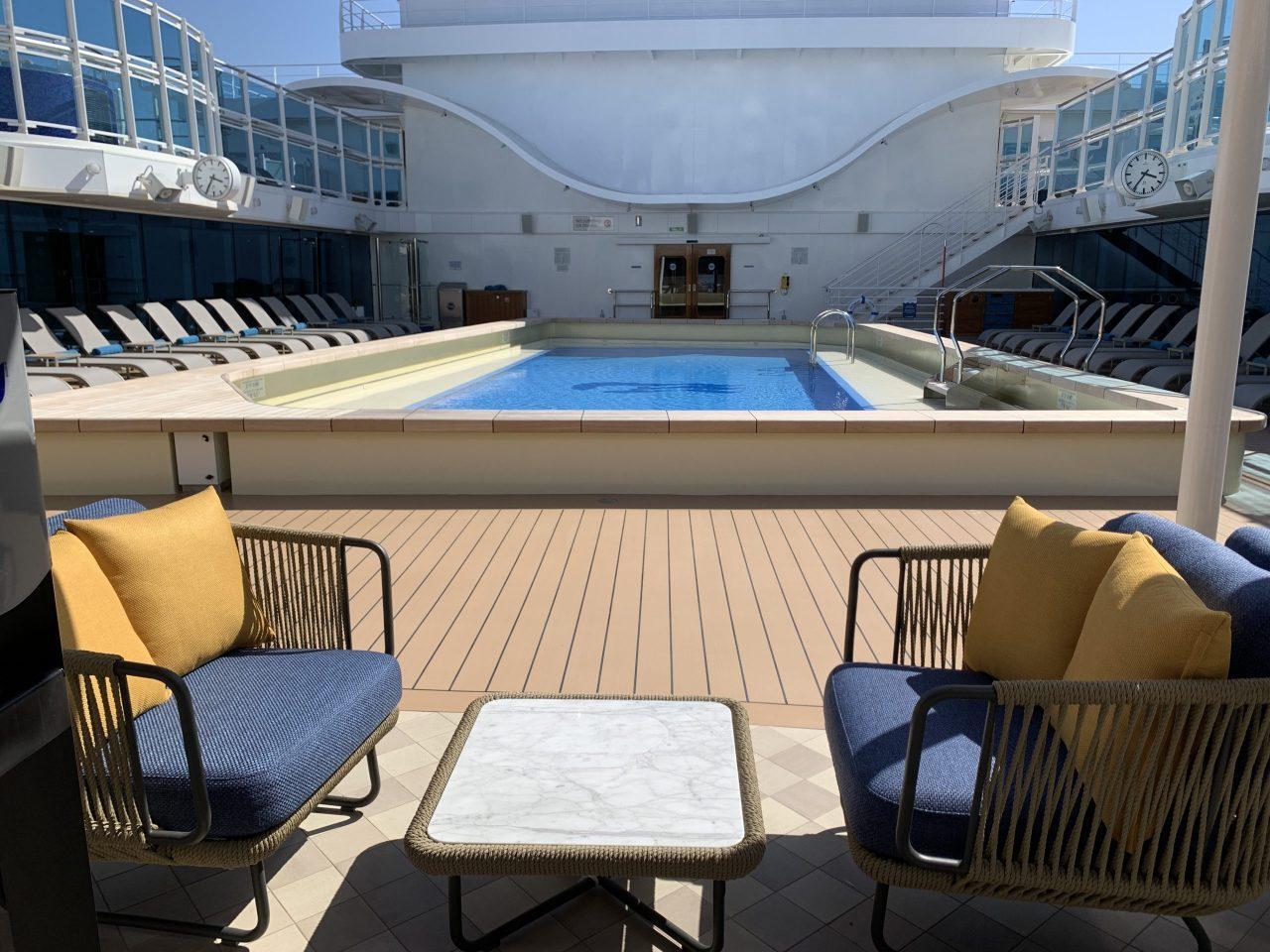 A cruise ship deck with a rectangular swimming pool surrounded by lounge chairs, viewed from a seating area with two blue chairs and a small table.