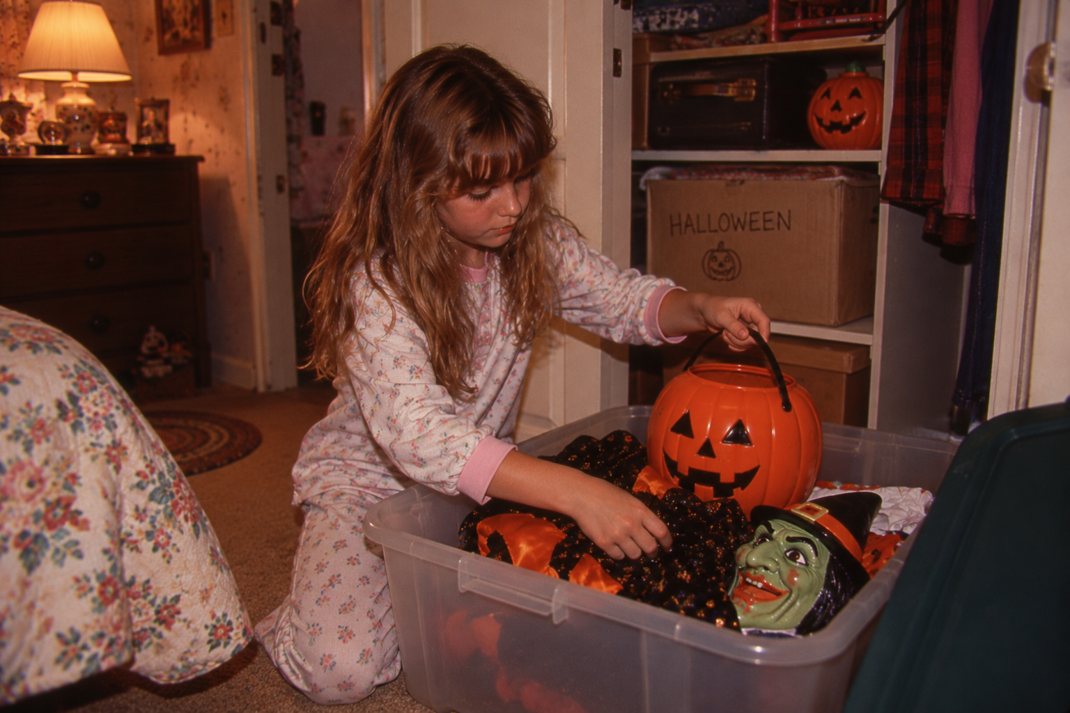 A young girl in pajamas kneeling on the floor, looking into a storage bin filled with Halloween decorations including a plastic jack-o’-lantern bucket and a witch mask.