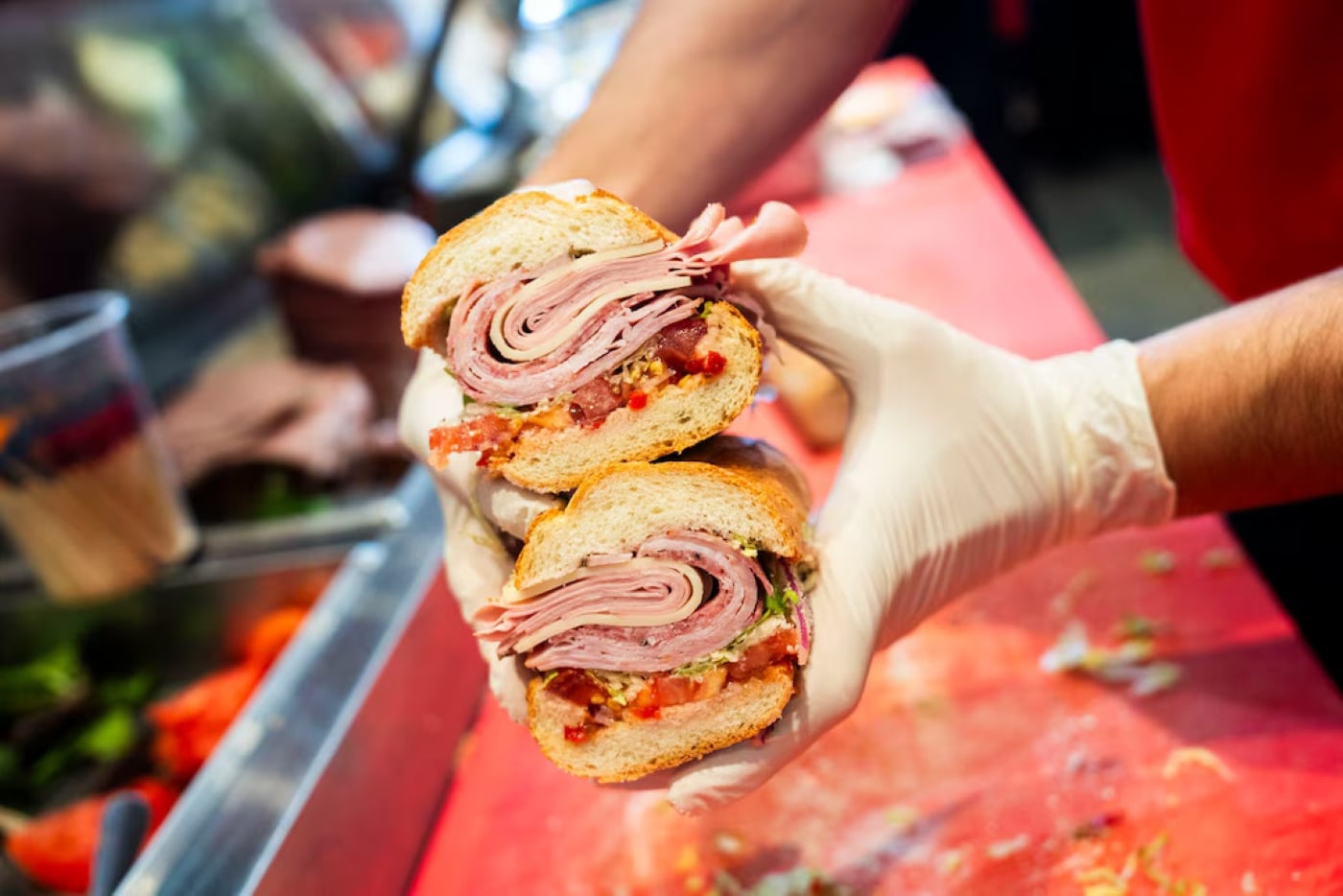 A person wearing a plastic glove holds a sliced deli sandwich stacked with multiple layers of ham, the kind often found at restaurants across the USA known for next-level hoagies.