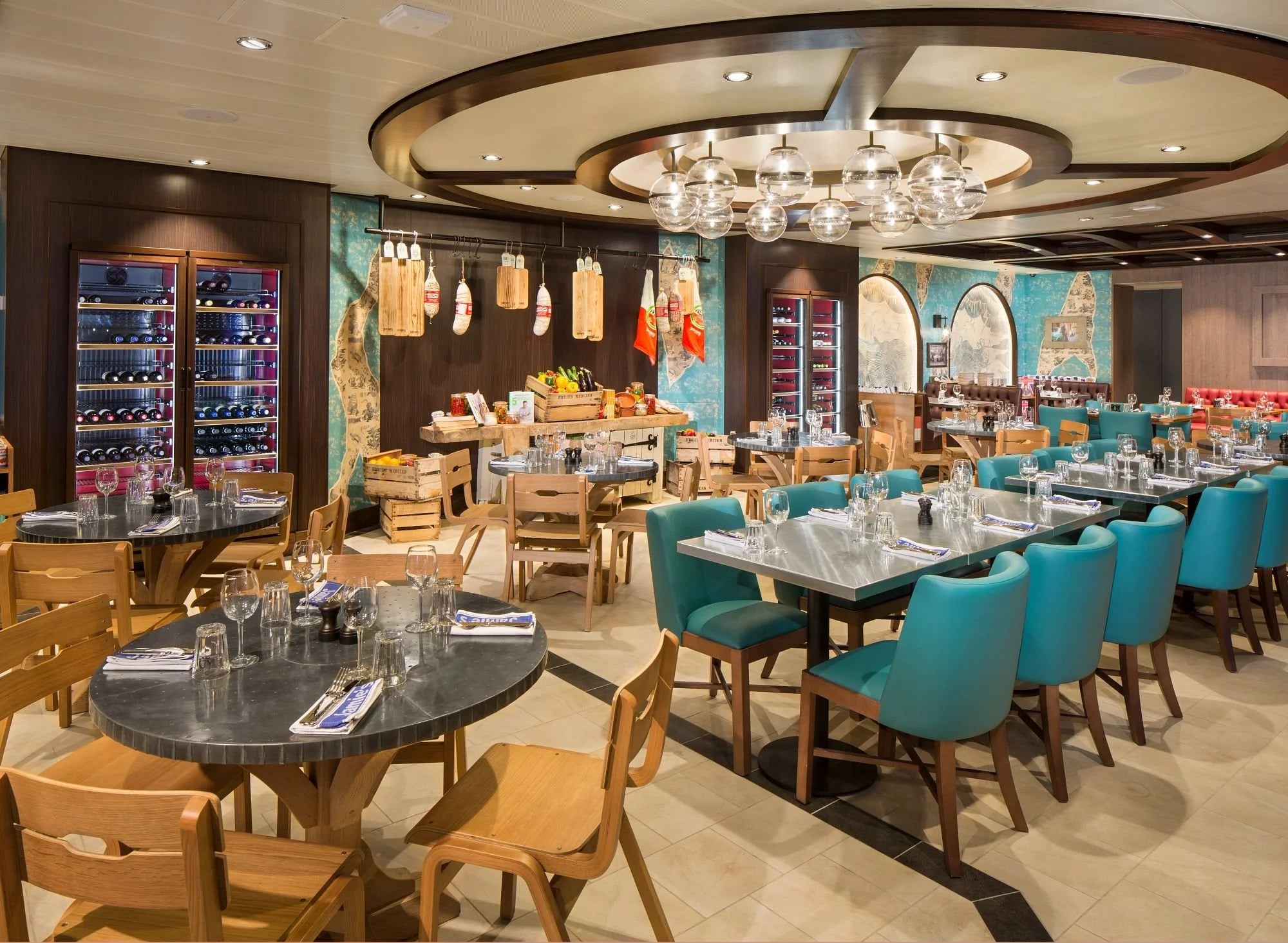 Interior of a restaurant with round and rectangular tables, teal chairs, and a display of hanging cured meats and cutting boards on the back wall.