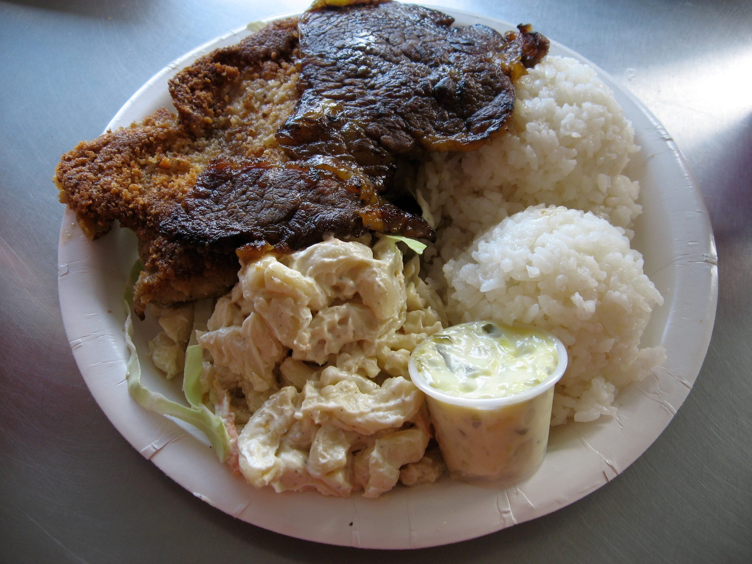A paper plate with a breaded cutlet, a piece of seared meat, two scoops of white rice, macaroni salad, and a small container of dipping sauce.