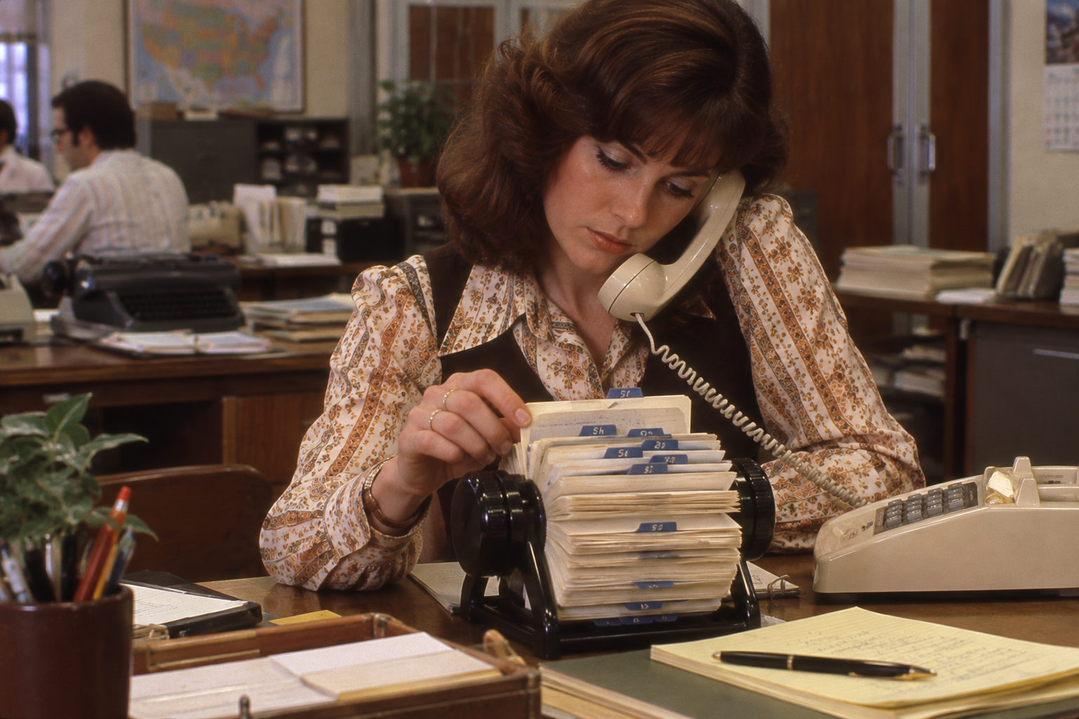 A woman at an office desk talks on a corded telephone while flipping through a rotary contact card file.