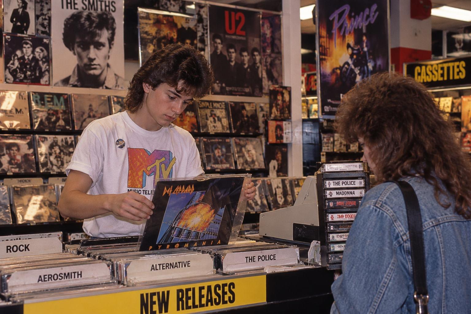 A young man in an MTV t-shirt shows a Def Leppard vinyl record to a woman in a denim jacket at a music store.