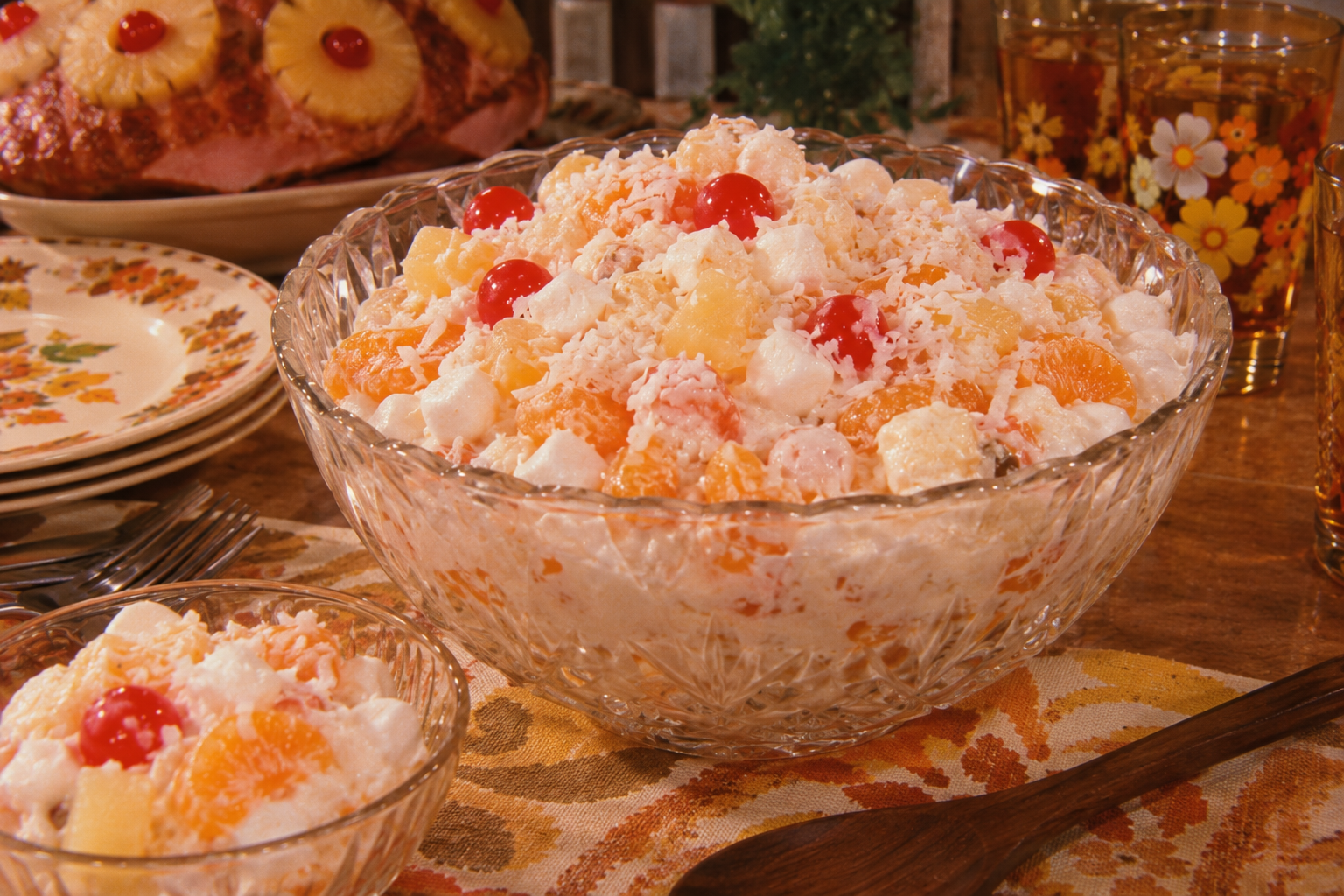 A glass bowl filled with ambrosia salad made of whipped cream, marshmallows, cherries, mandarin oranges, and pineapple sits on a table with vintage dishes and glasses with floral designs.