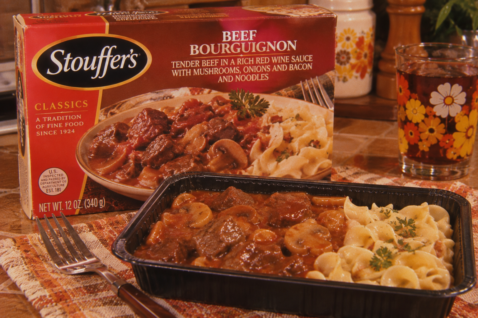 A tray of Stouffer’s Beef Bourguignon with noodles sits on a placemat next to a fork, knife, and a glass of iced tea. The product box featuring the same meal is in the background. 1980s foods.