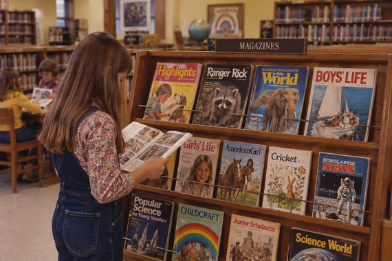 A young girl stands in a library, reading a magazine in front of a rack filled with various children’s magazines.