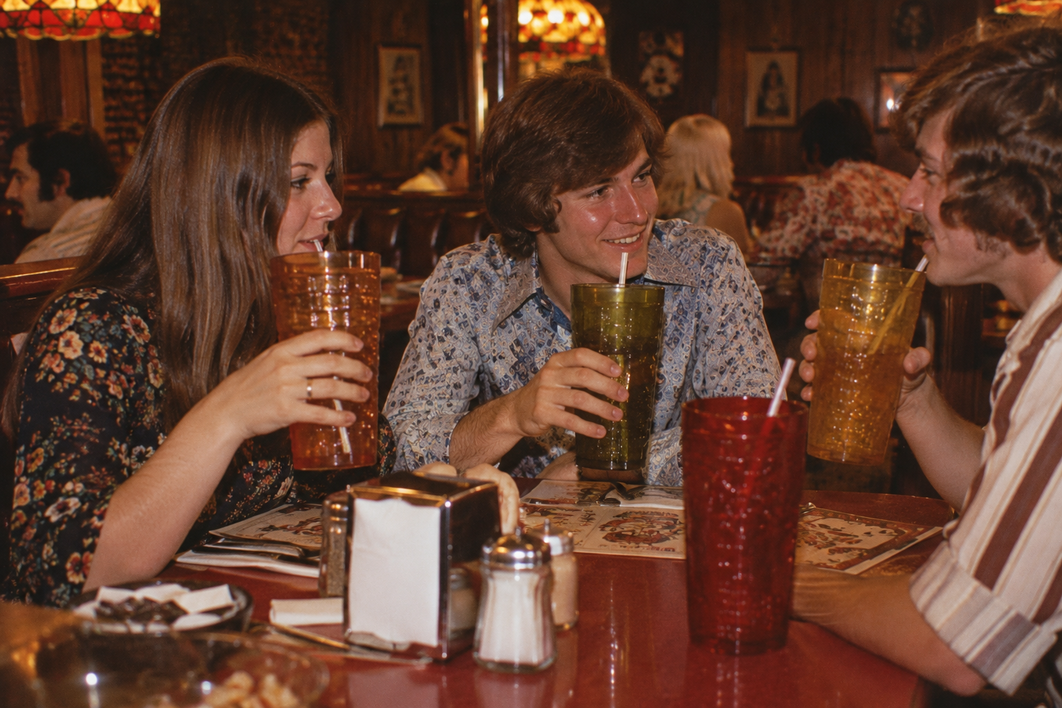 Three young adults sit at a restaurant table, smiling and drinking from large colorful cups with straws. Menus, a napkin holder, and salt and pepper shakers are on the table. The setting has warm lighting and wood-paneled walls.
