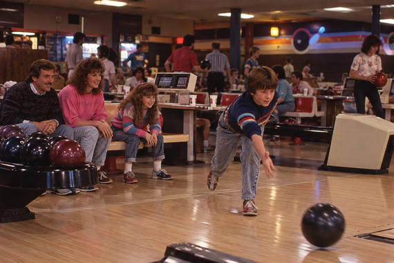 A young boy bowls a ball down a lane while a family watches from the seating area at a bowling alley.