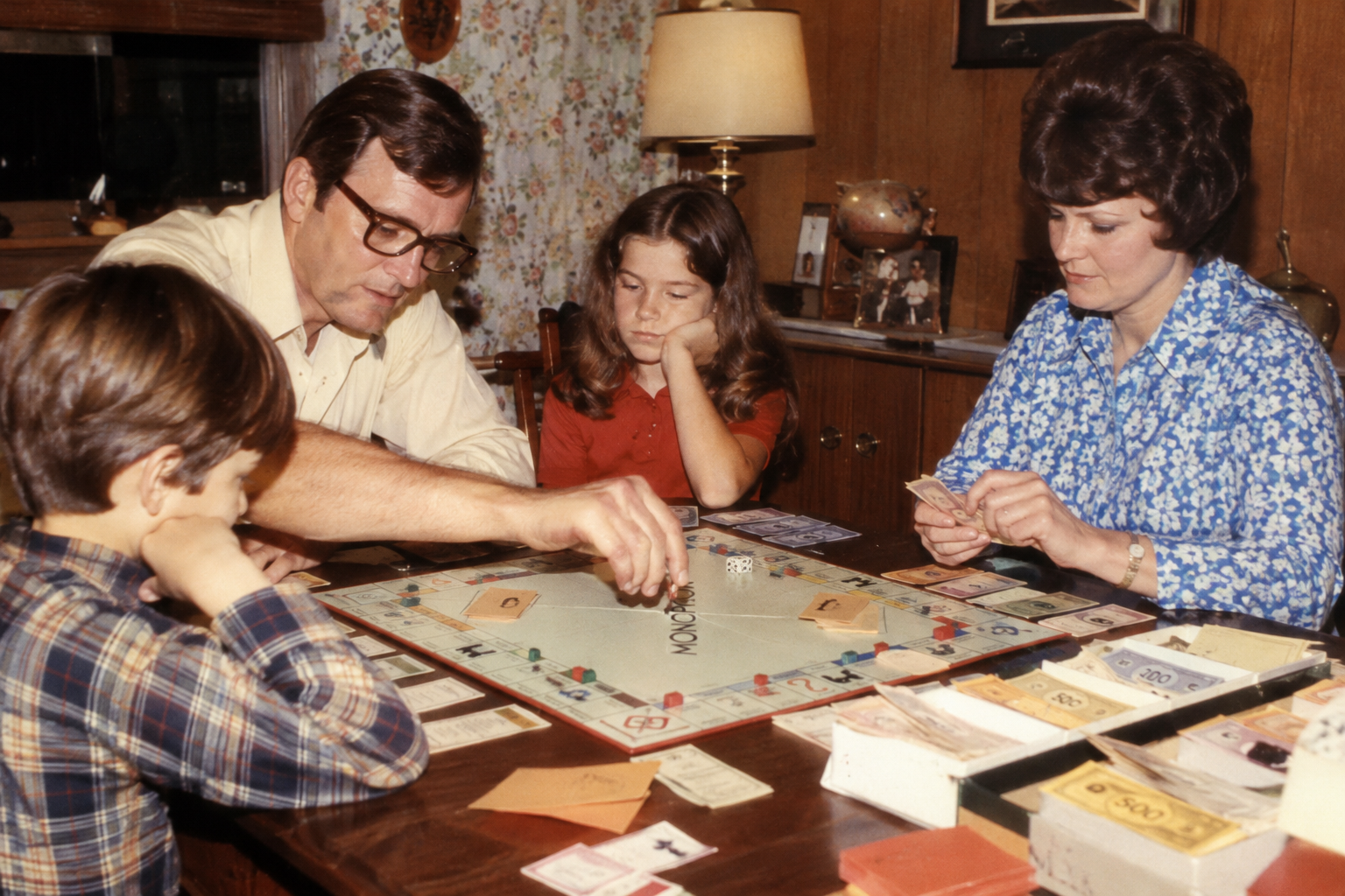 A family of four playing a game of Monopoly around a wooden table.