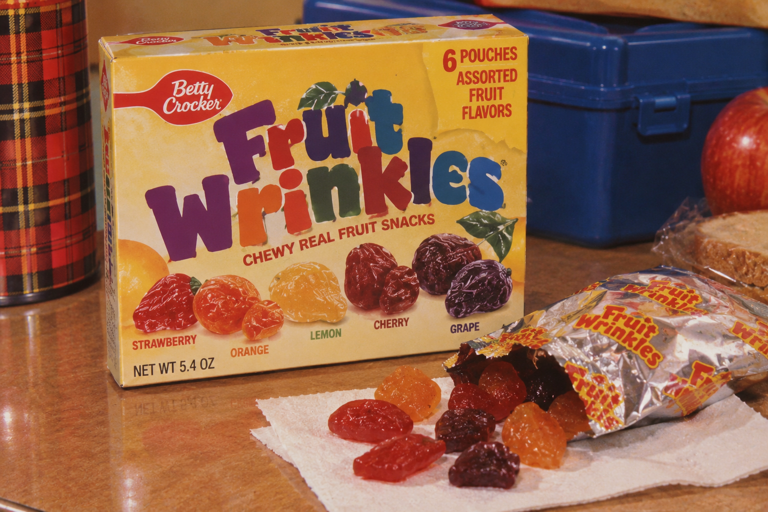 A box of Betty Crocker Fruit Wrinkles fruit snacks sits on a table, with an open pouch and several colorful fruit snacks displayed on a napkin in front of it.