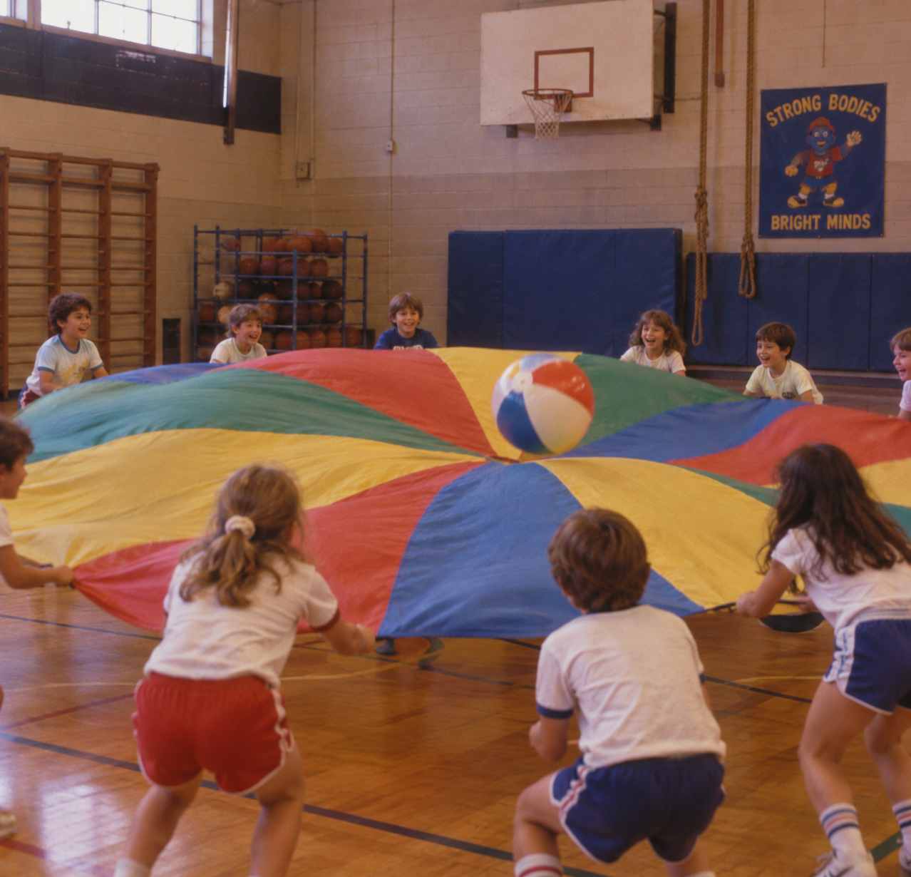 A group of children in a gym playing with a large, multi-colored circular parachute with a beach ball bouncing on top.