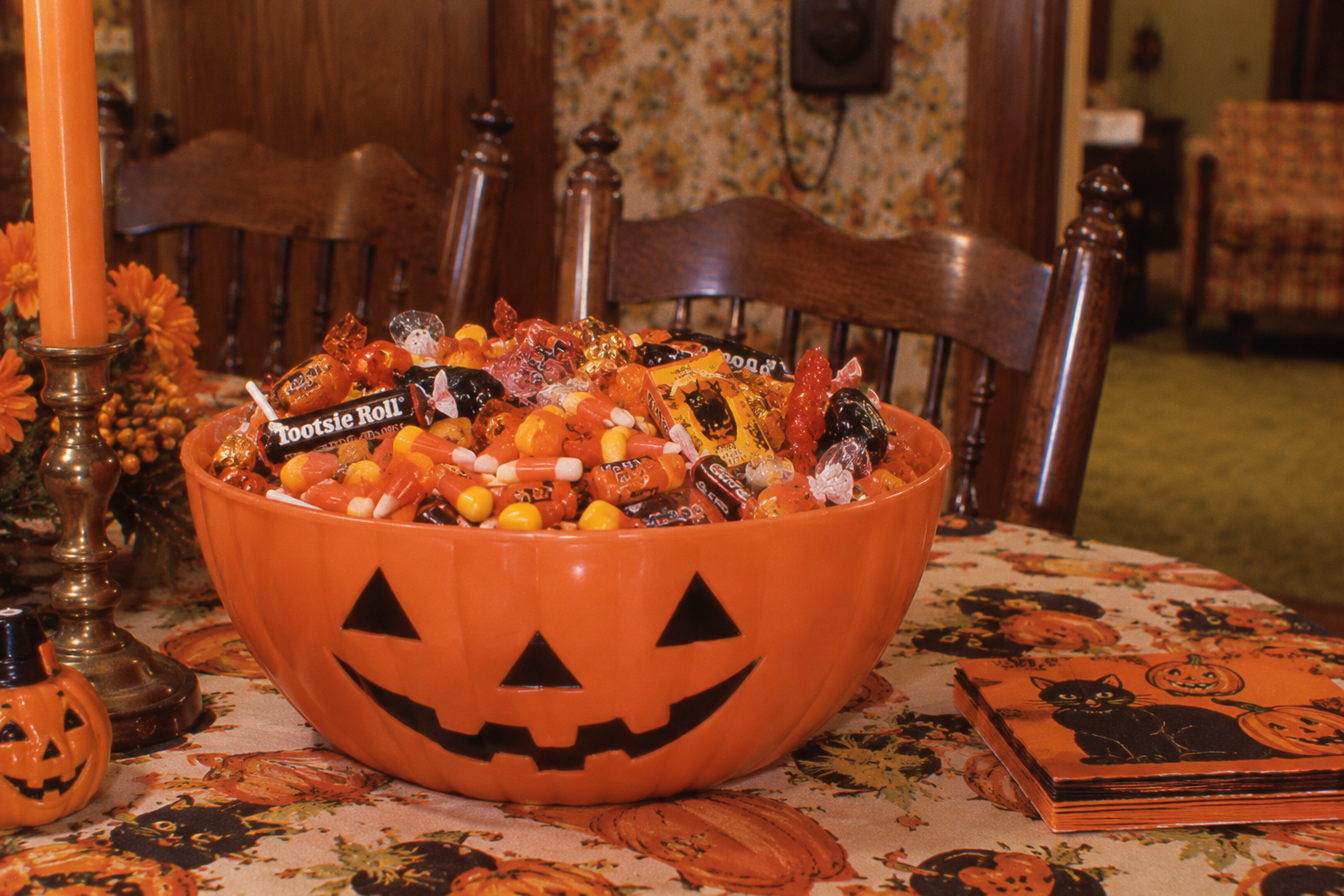 An orange jack-o’-lantern bowl filled with assorted Halloween candy sits on a table covered in a fall-themed tablecloth.