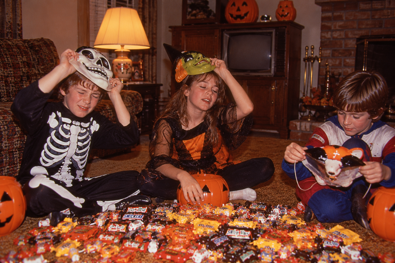 Three children in Halloween costumes sit on a living room floor surrounded by a large pile of candy.