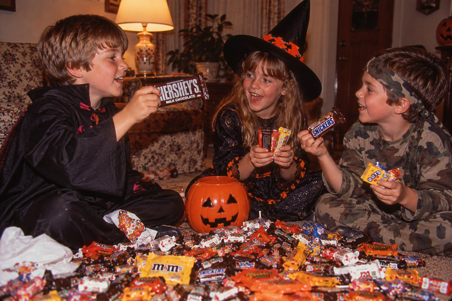 Three children in Halloween costumes sit on the floor surrounded by a large pile of candy, holding up various candy bars.