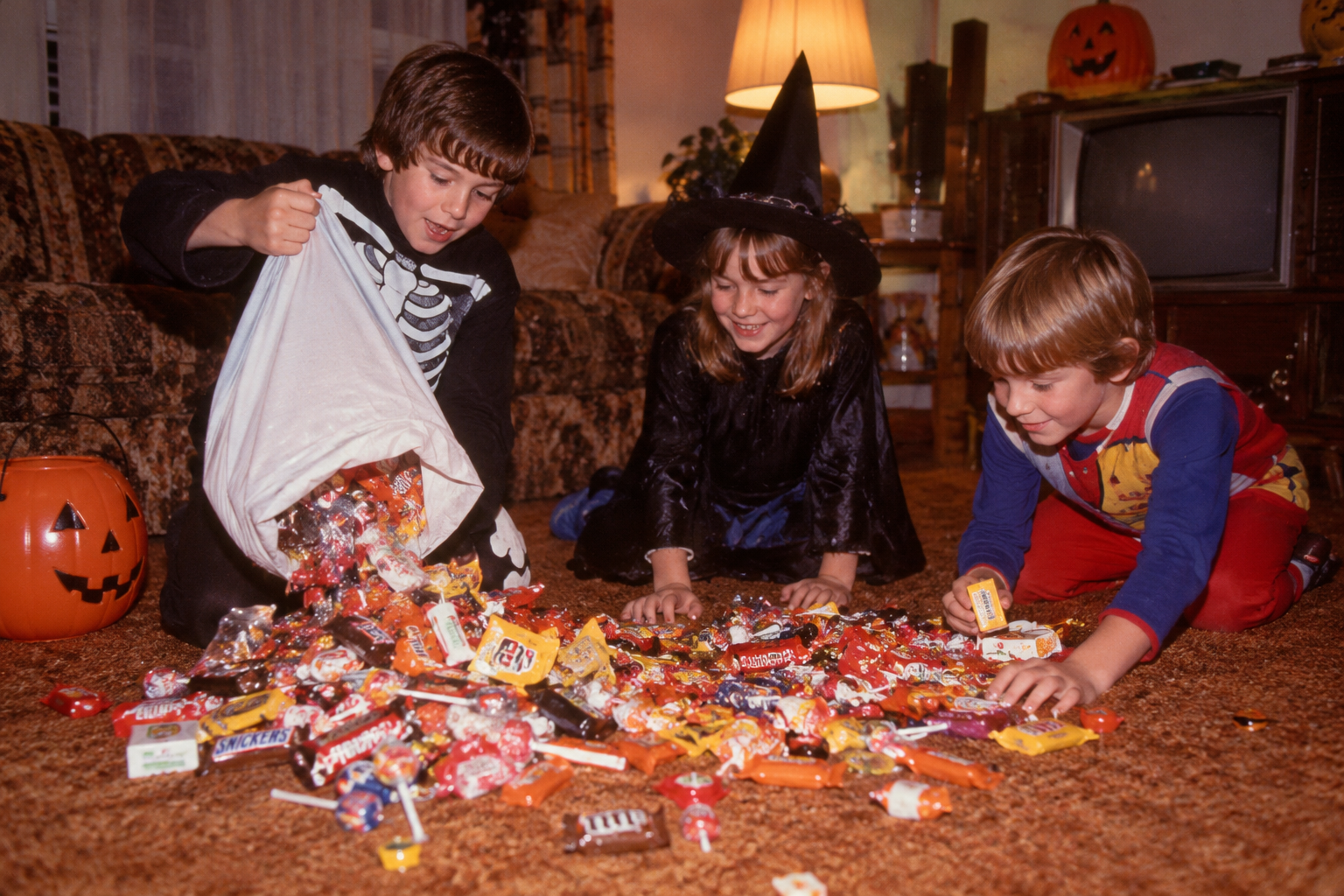 Three children in Halloween costumes sit on a carpet, sorting through a large pile of candy.