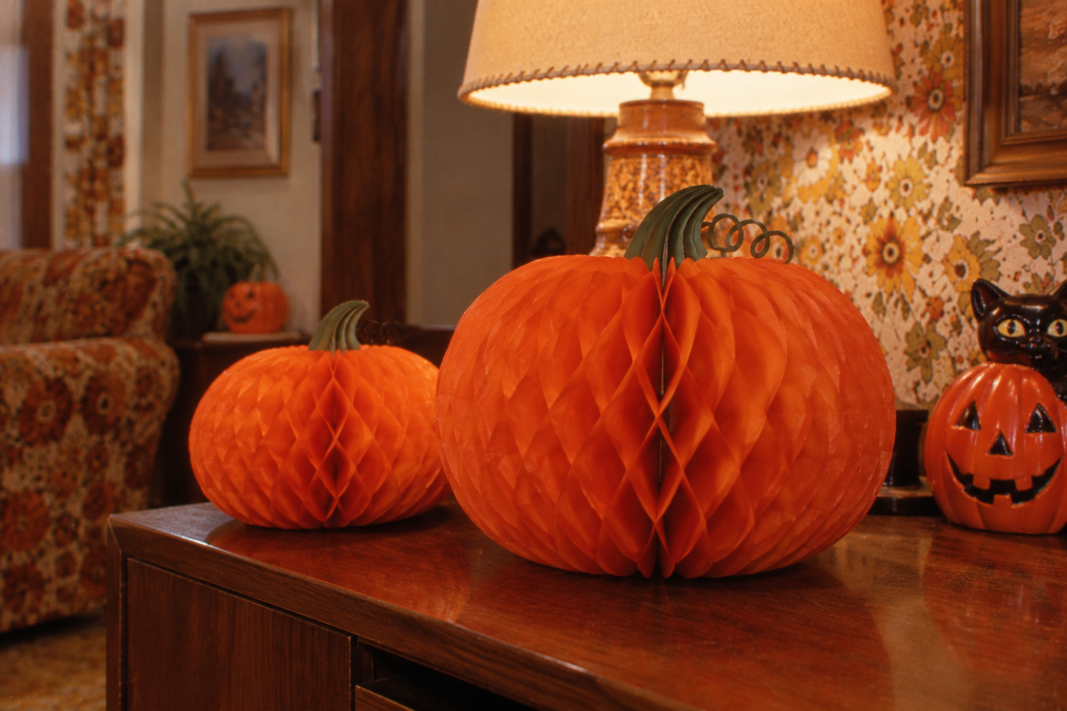Two orange honeycomb paper pumpkins sitting on a wooden sideboard next to a ceramic jack-o’-lantern and a table lamp.