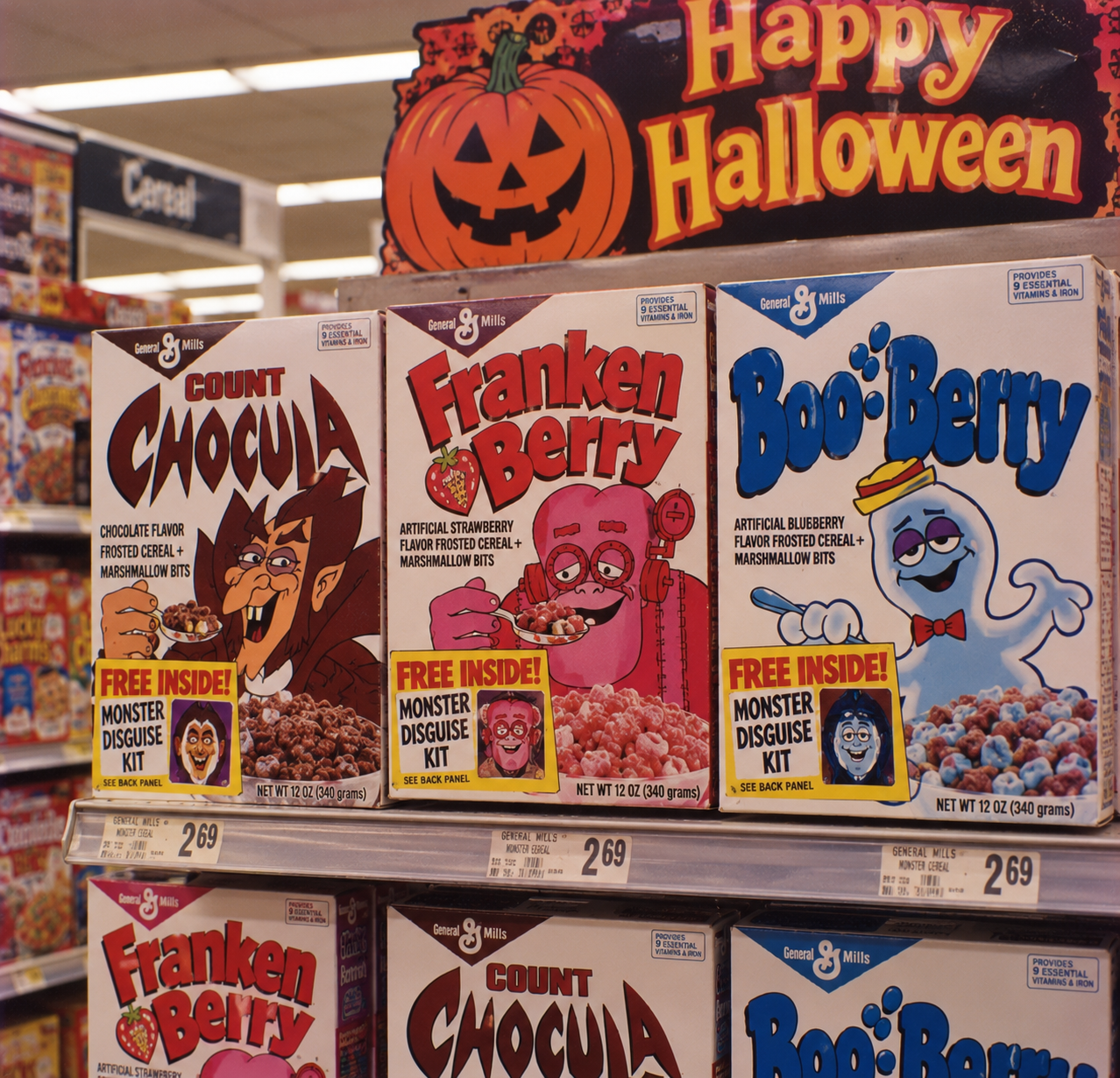 Three boxes of General Mills monster cereal—Count Chocula, Franken Berry, and Boo Berry—on a grocery store shelf under a “Happy Halloween” sign with a jack-o’-lantern illustration.