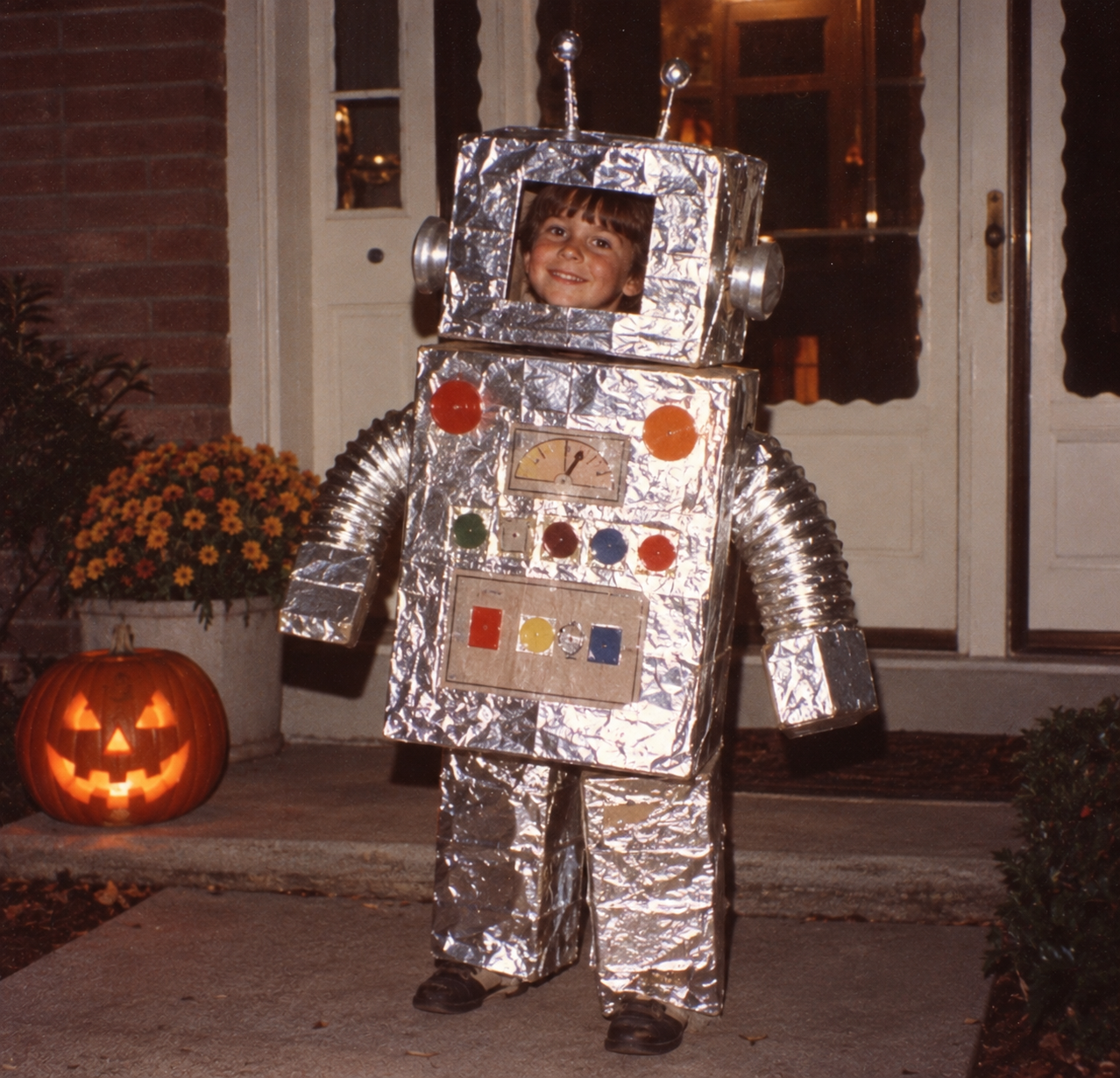 A child in a homemade foil robot costume stands on a porch next to a lit jack-o’-lantern.