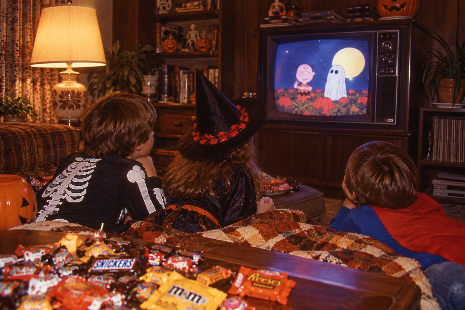 Three children in Halloween costumes lie on a floor watching “It’s the Great Pumpkin, Charlie Brown” on a television, with a large pile of candy on a coffee table in the foreground.