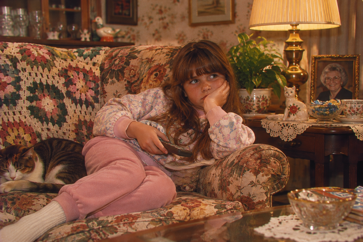 A young girl sits on a floral sofa with a cat, holding a remote control and resting her head on her hand.