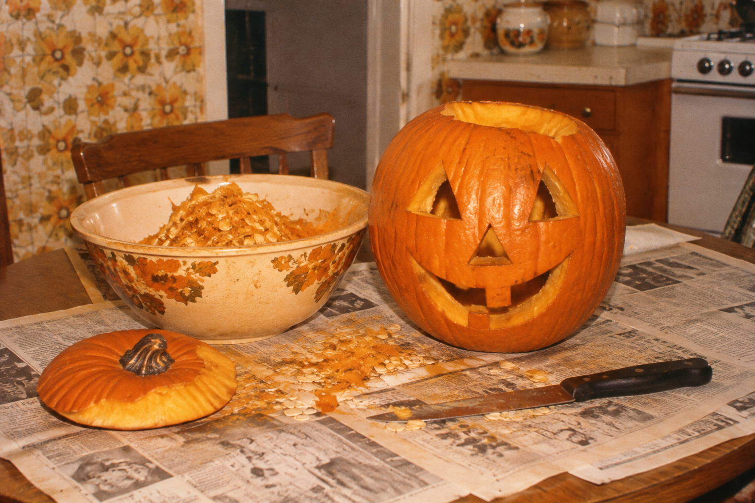 A carved jack-o’-lantern, its lid, and a bowl of pumpkin seeds and pulp sitting on a table covered in newspaper.