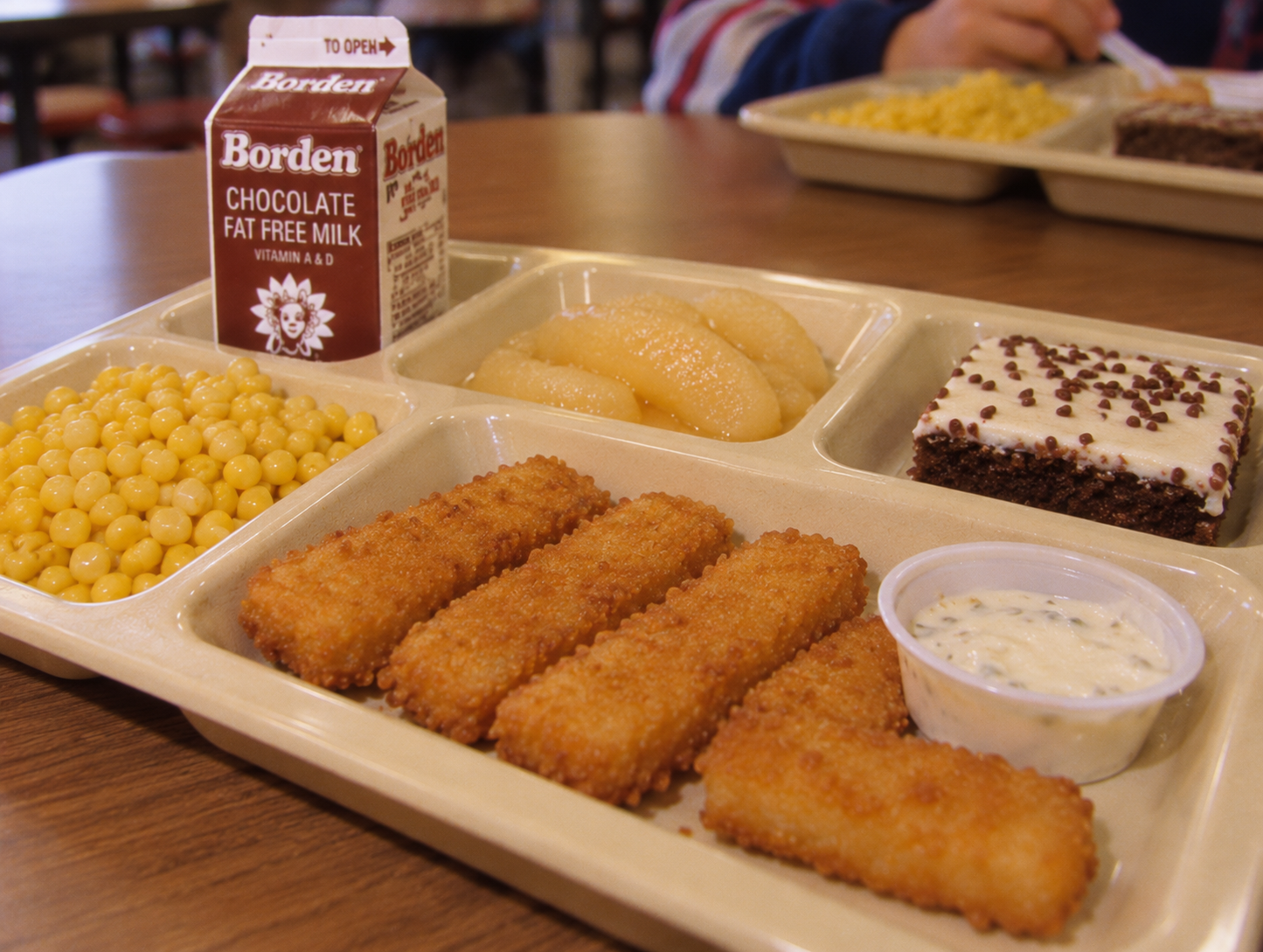 A school lunch tray with breaded fish sticks, tartar sauce, corn, applesauce, a chocolate brownie, and a carton of Borden chocolate milk.