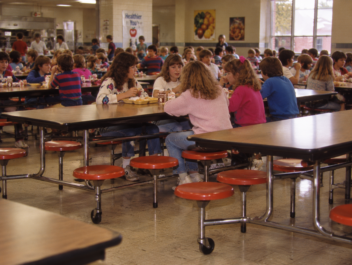Students eating lunch at tables in a school cafeteria. 1980s school cafeteria.