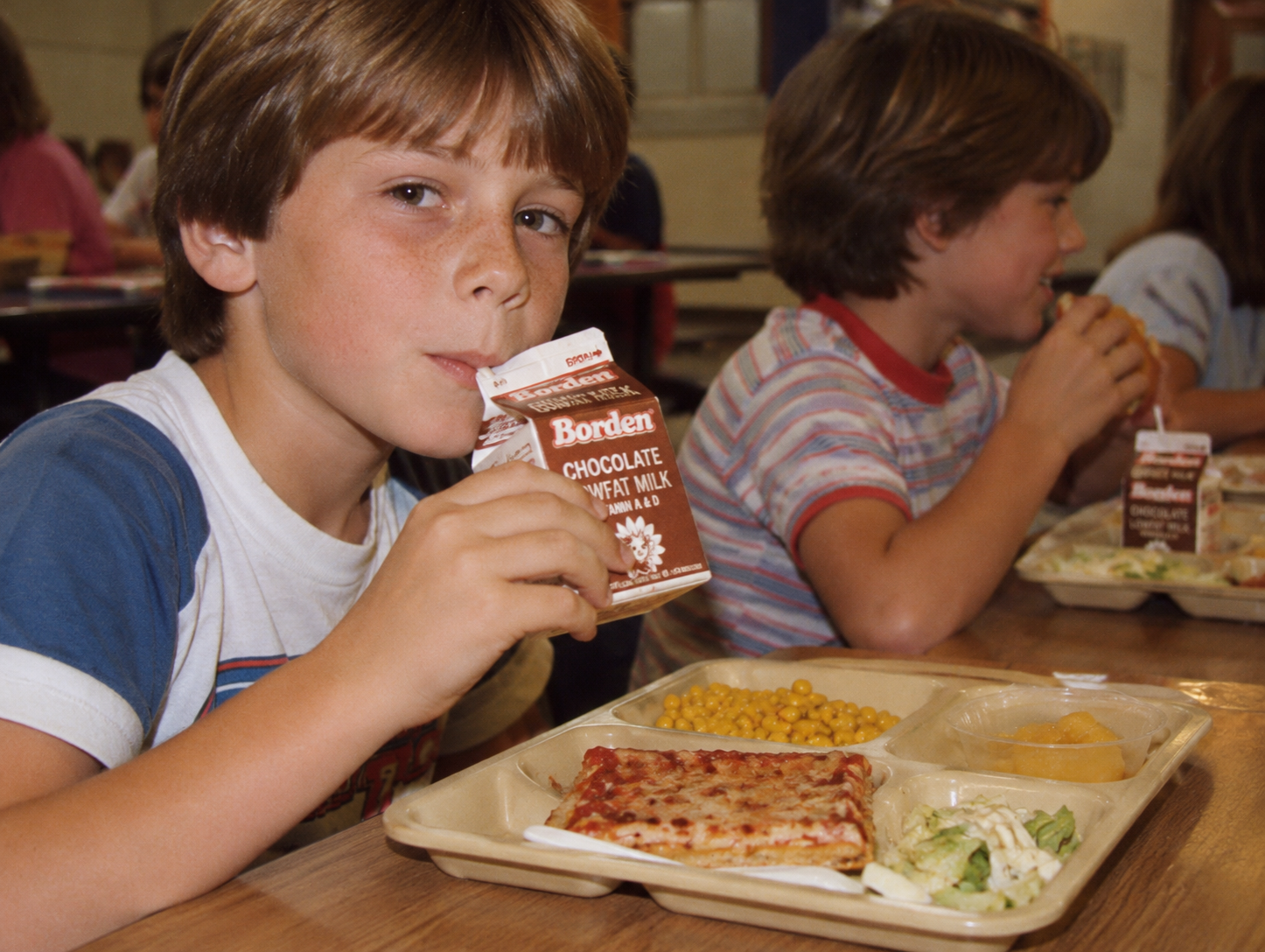 A boy at a school cafeteria table drinks from a carton of Borden chocolate milk while looking toward the camera, with a school lunch tray in front of him.