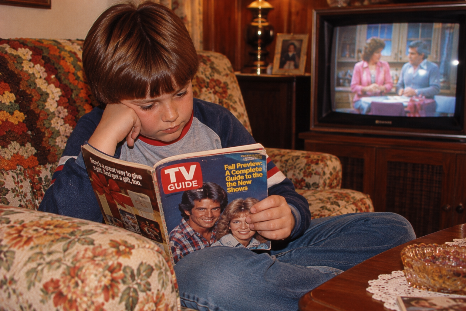 A young boy sitting on a floral couch reads a TV Guide magazine while a television plays in the background.