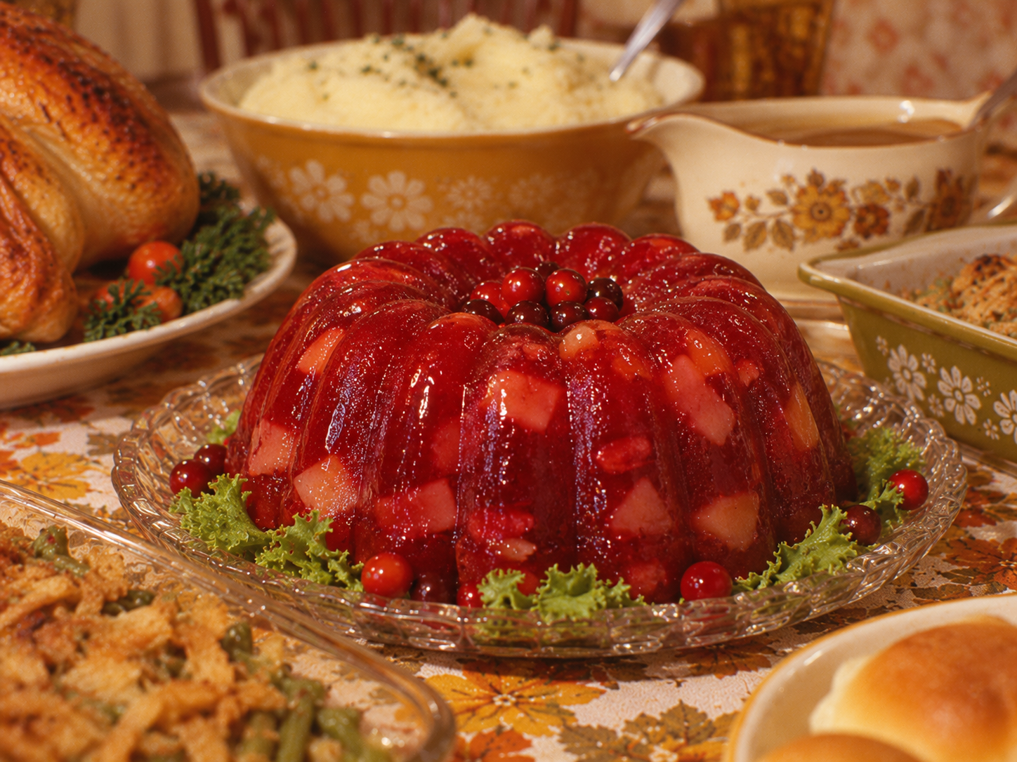 A gelatinous cranberry mold filled with fruit pieces sits on a glass platter, surrounded by Thanksgiving side dishes like turkey, mashed potatoes, and gravy.
