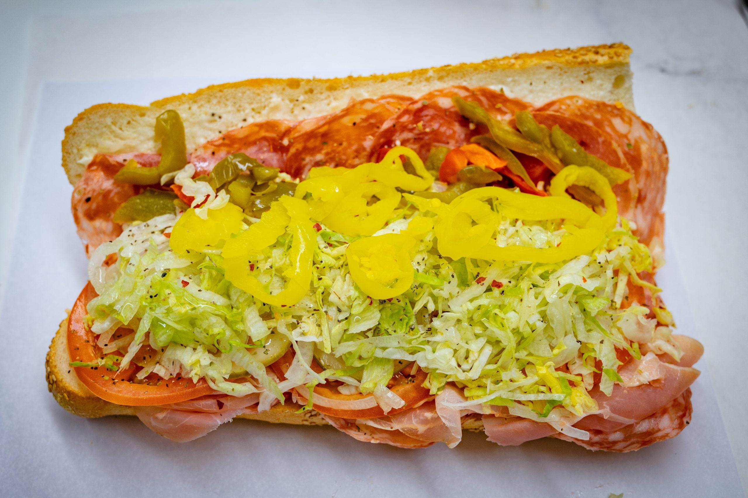 A sub sandwich on a crusty roll filled with sliced deli meats, shredded lettuce, tomato slices, and yellow banana peppers. Restaurants Across The USA Known For Next-Level Hoagies