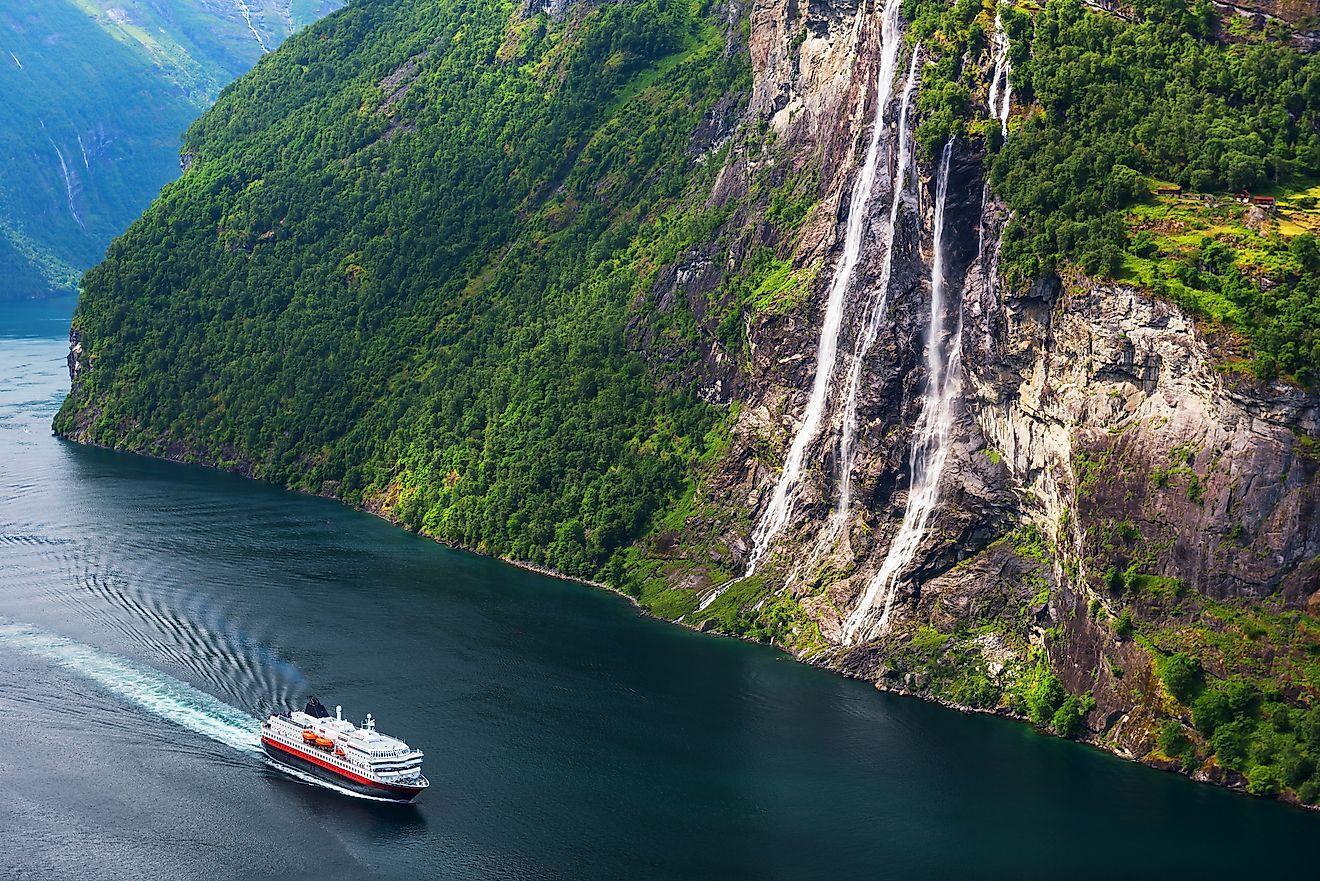 A cruise ship sails through a deep fjord past a tall waterfall cascading down a steep, forested mountain.