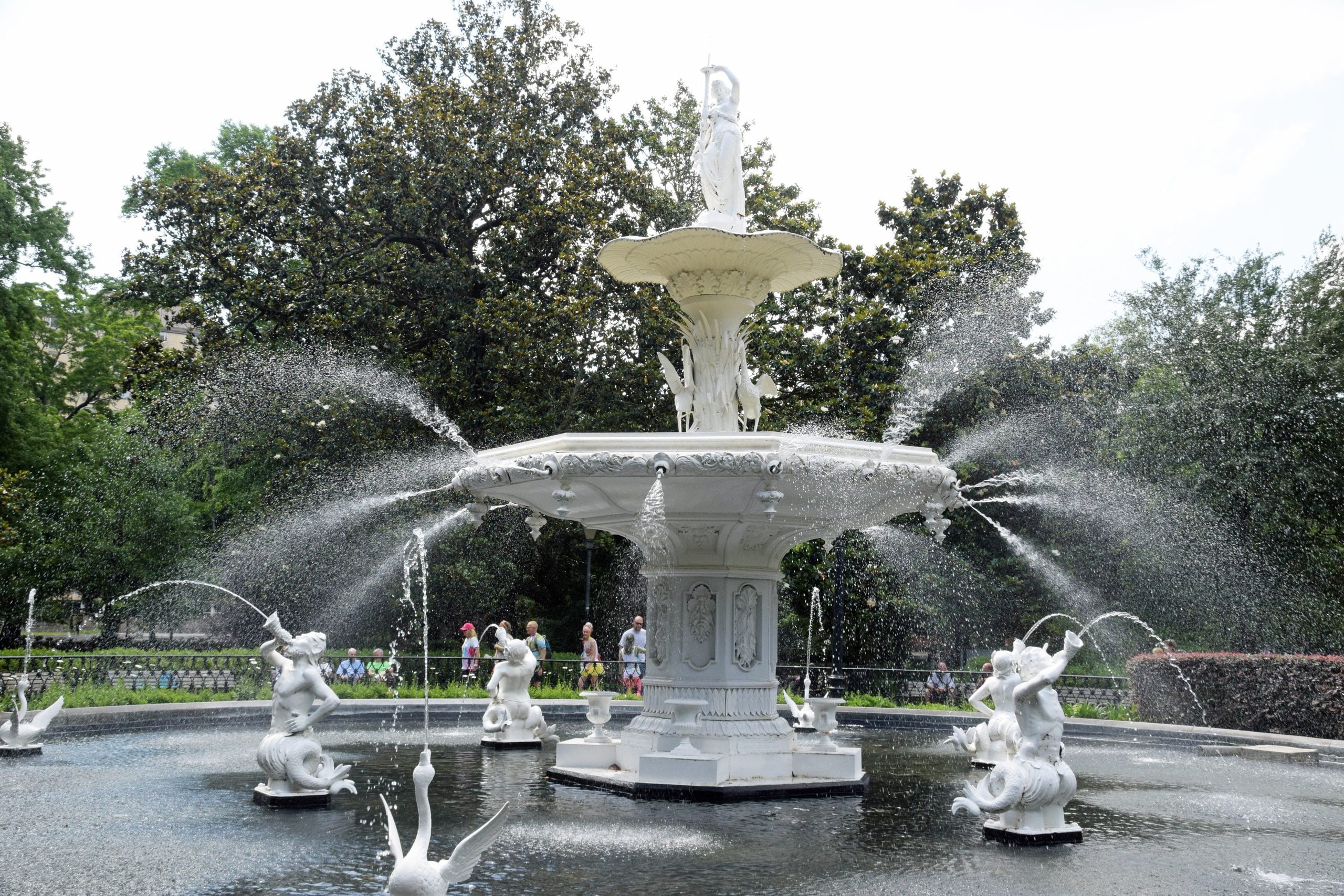 A large, ornate white fountain with multiple tiers and statues, spraying water in arcs, stands in a park surrounded by greenery and people.