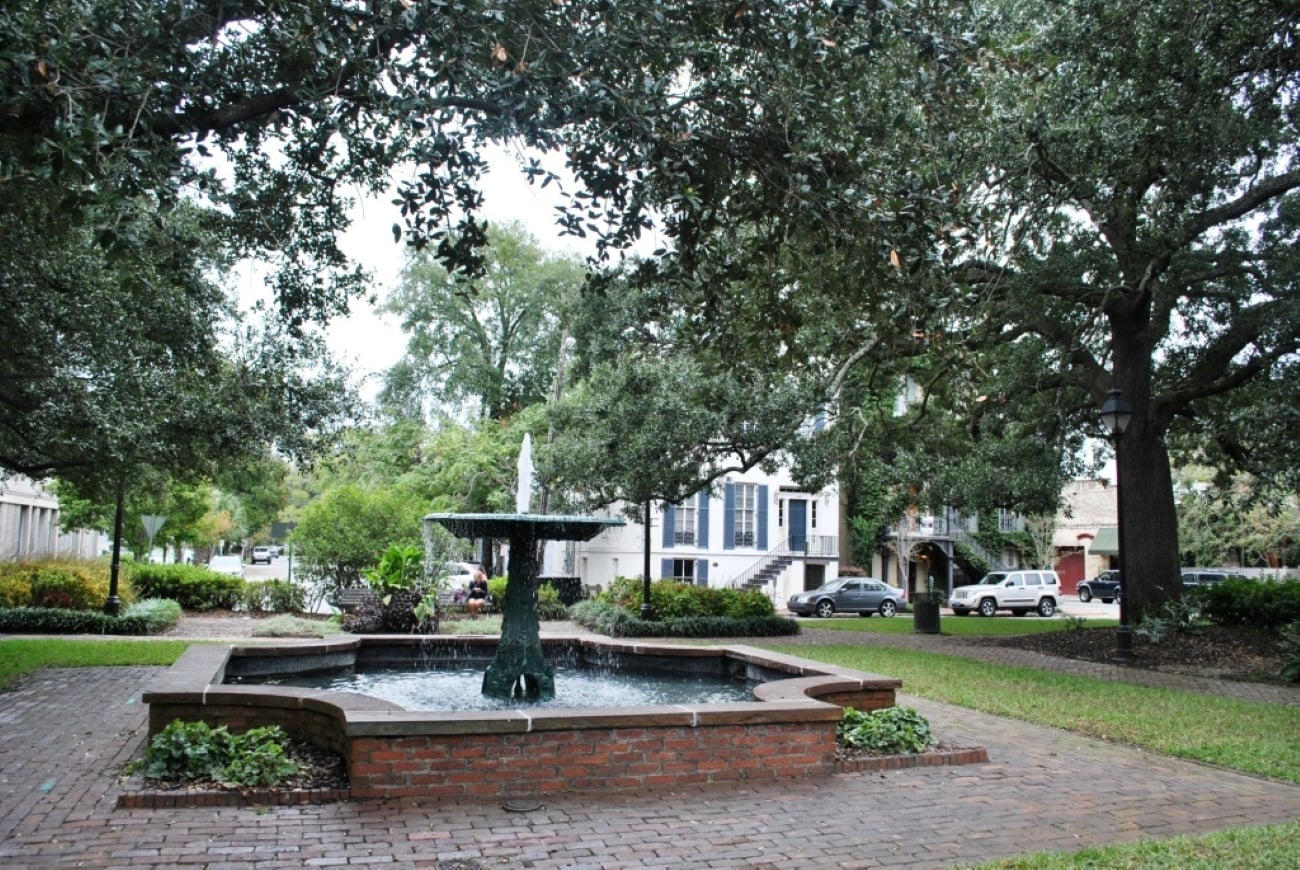 A tiered stone fountain sits in the center of a brick courtyard, surrounded by large oak trees and nearby buildings.