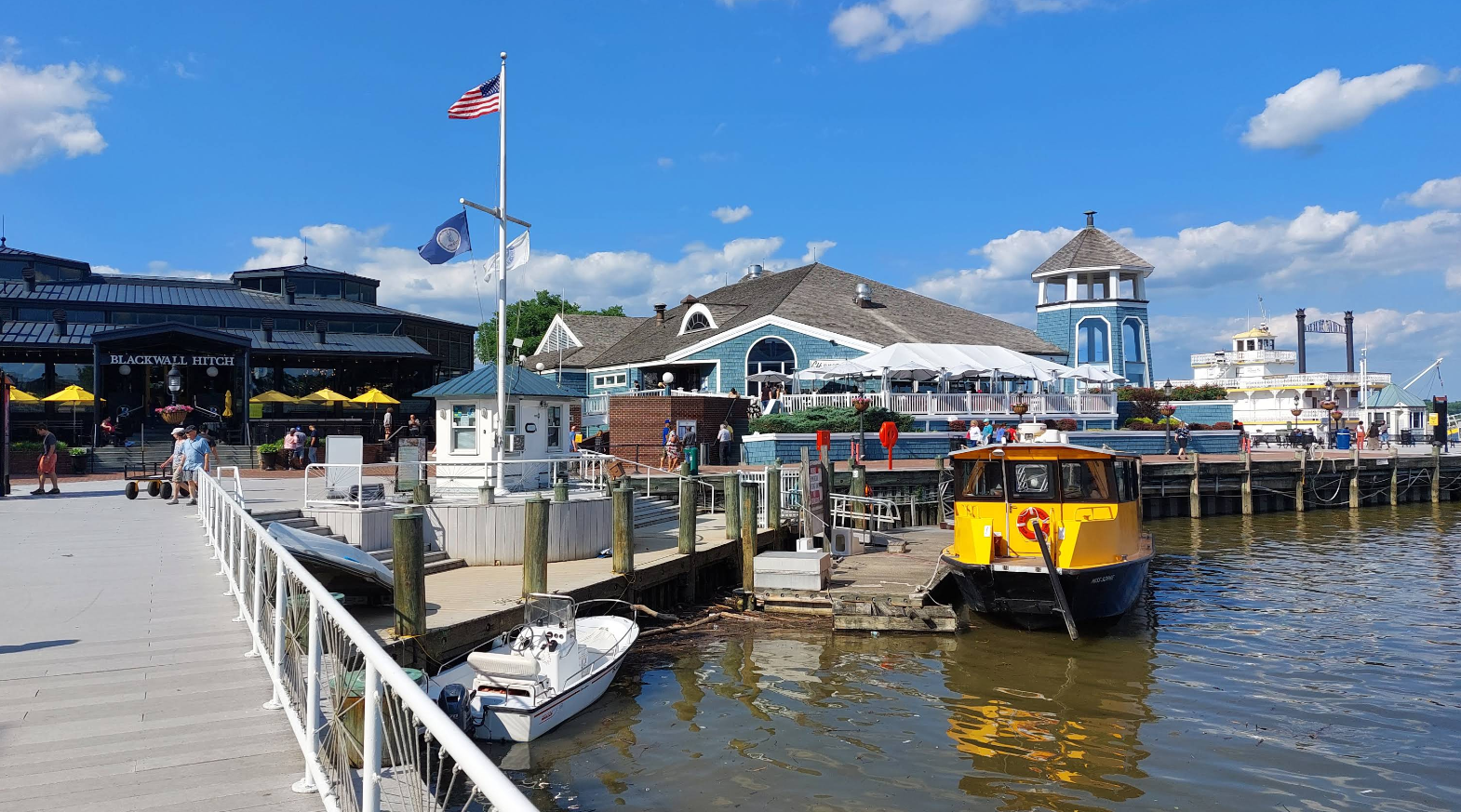 A small boat and a yellow water taxi docked at a waterfront pier, where people stroll by—perfect for trips where one great neighborhood meets blue buildings and an American flag under a bright, cloud-scattered sky.