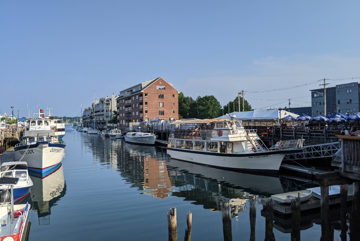 Boats are docked along a calm waterfront in one great neighborhood, lined with brick and wooden buildings, their reflections shimmering on a sunny day under a clear blue sky—the perfect spot for scenic trips.