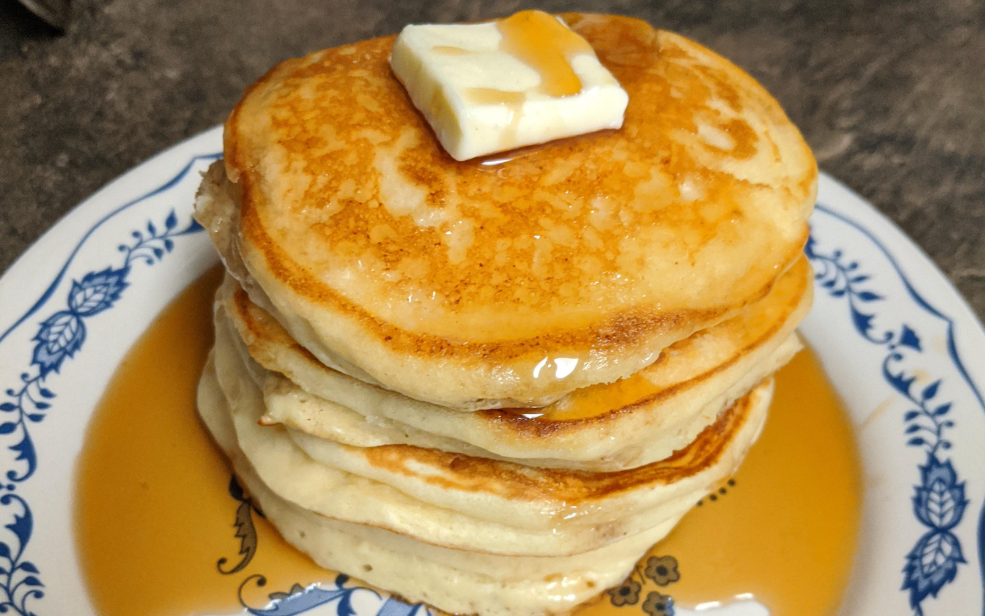 A stack of pancakes, perfect for trying new pancake recipes, topped with a pat of butter and drizzled with syrup on a patterned plate.
