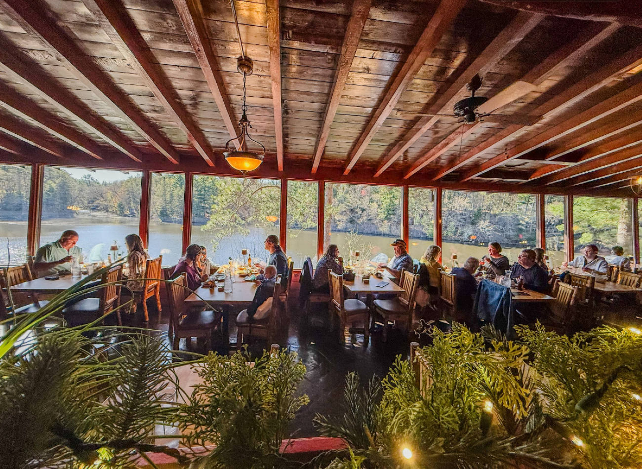 People dining at tables in a rustic, wood-paneled restaurant with large windows overlooking a lake.