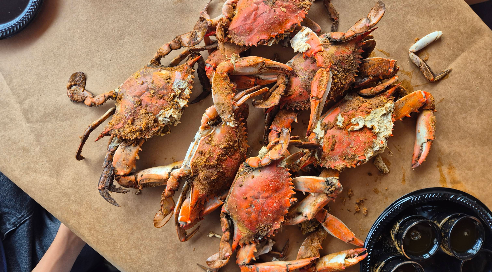 A pile of steamed blue crabs seasoned with spice, served on brown butcher paper next to small containers of dipping sauce.