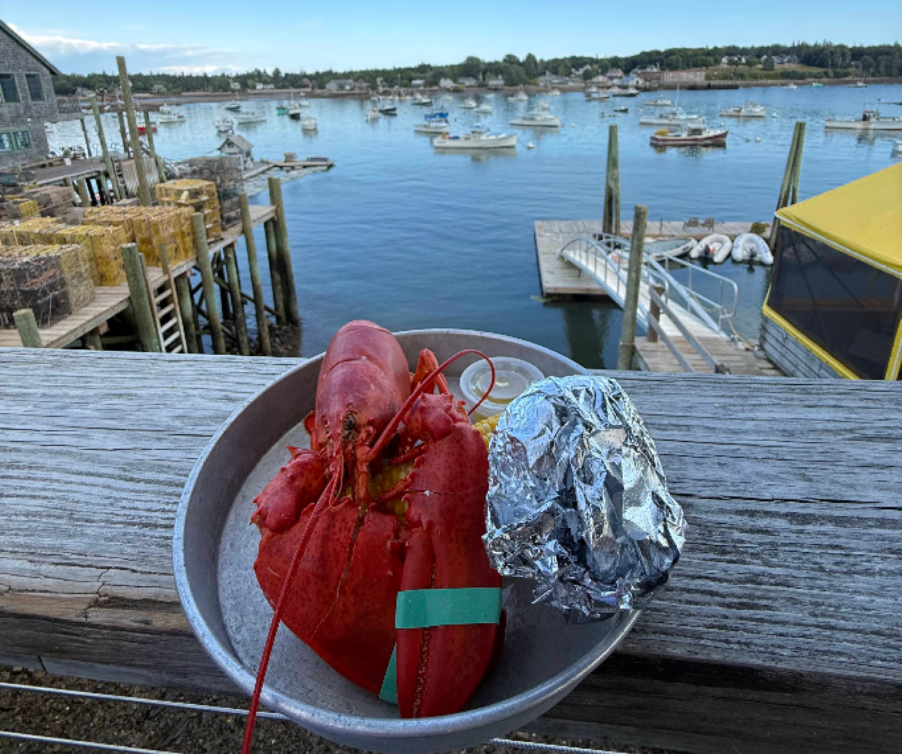 A whole cooked lobster, corn on the cob, and a foil-wrapped item on a metal plate, set against a backdrop of a harbor with boats and a dock.