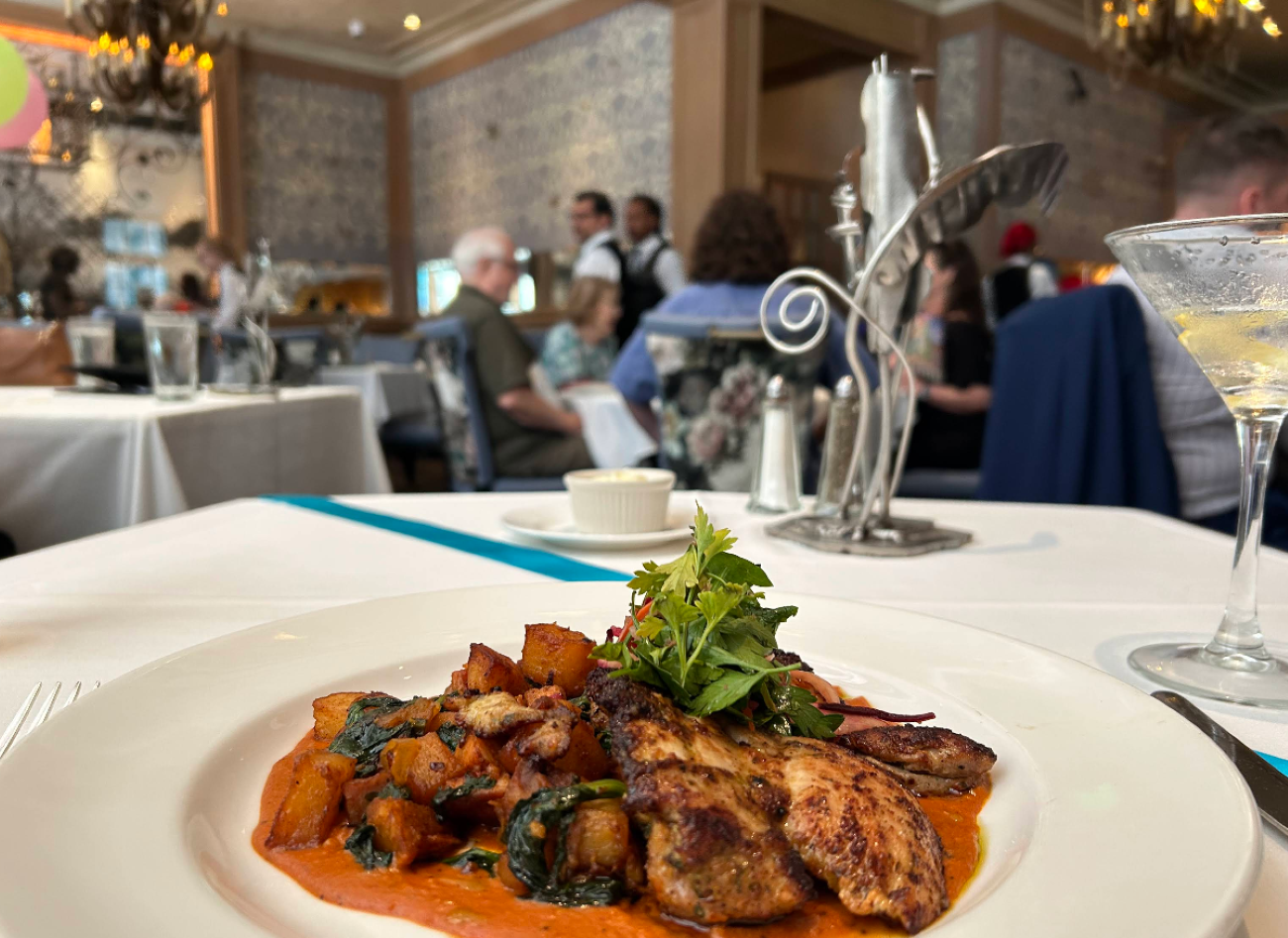A plate of roasted chicken and sautéed potatoes with greens in a red sauce, served in a restaurant.