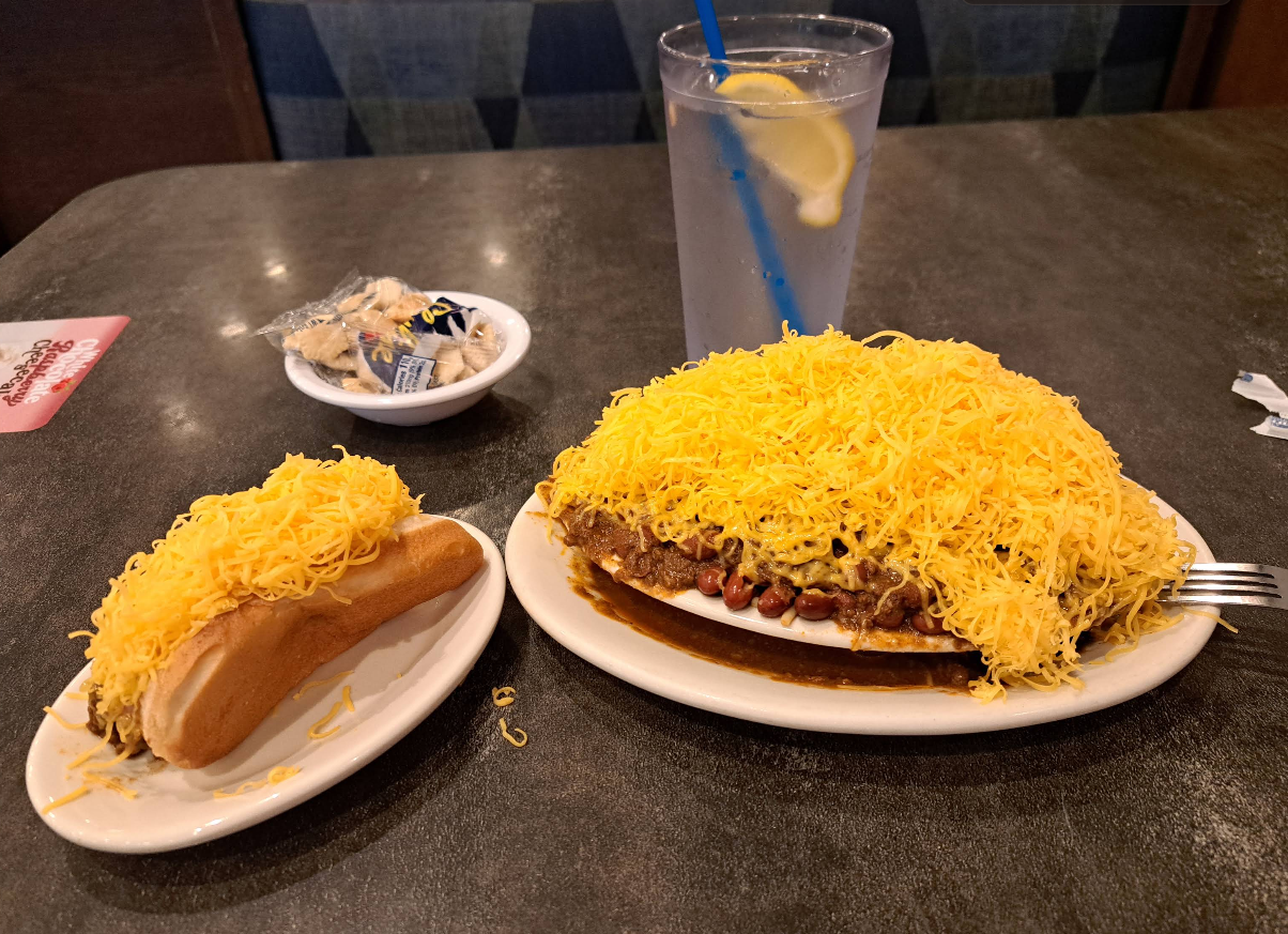A chili cheese dog and a plate of chili beans covered in shredded yellow cheese, served with a glass of water and crackers.