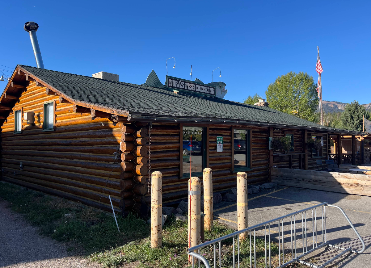 A log cabin building housing Nora’s Fish Creek Inn, featuring a large fish sign on the roof and a parking area in front.