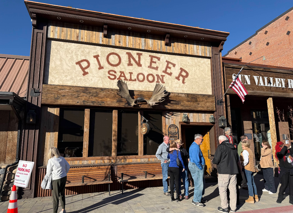 A group of people standing outside the Pioneer Saloon in front of a building with moose antlers mounted above the entrance.