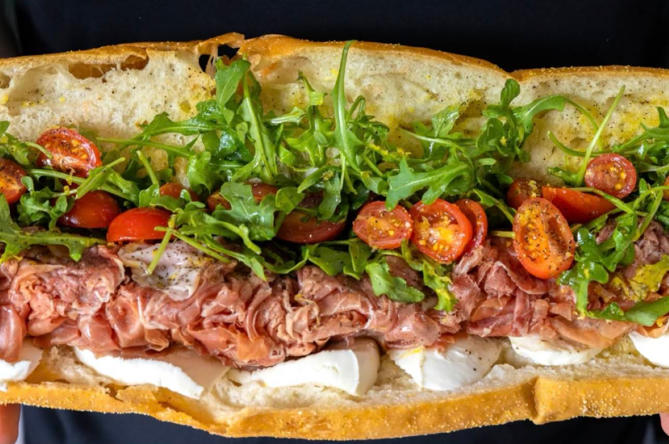 A roast beef sandwich on a crusty roll with fresh mozzarella, arugula, and halved cherry tomatoes, typical of restaurants across the USA known for next-level hoagies.