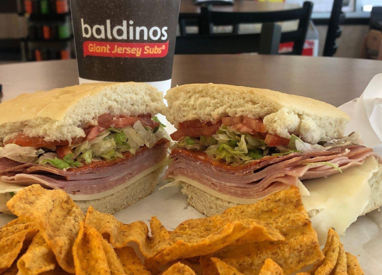 A halved sub sandwich with deli meat, cheese, lettuce, and tomato, served with corn chips in front of a Baldinos Giant Jersey Subs cup, representing restaurants across the USA known for next-level hoagies.