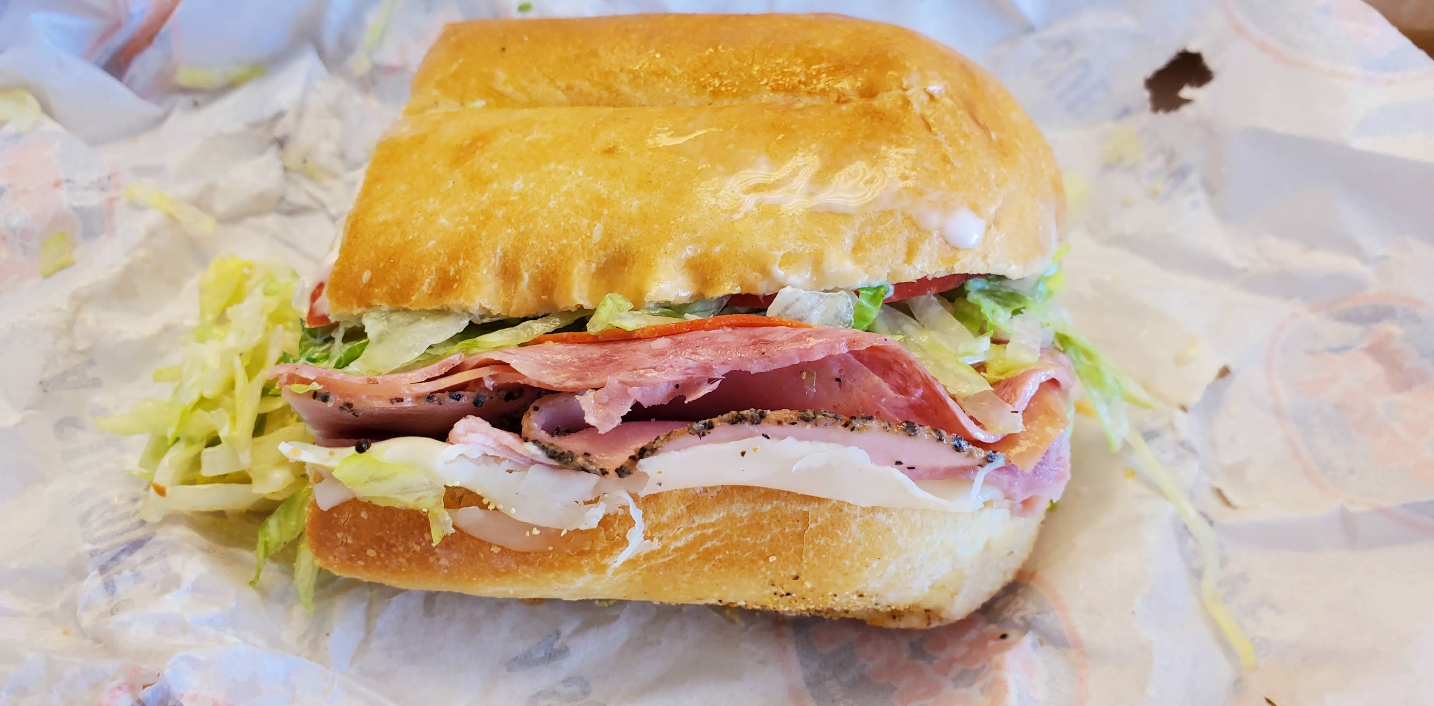A deli sandwich with sliced meat, lettuce, and tomato on a crusty roll, sitting on sandwich wrap paper like those found at restaurants across the USA known for next-level hoagies.