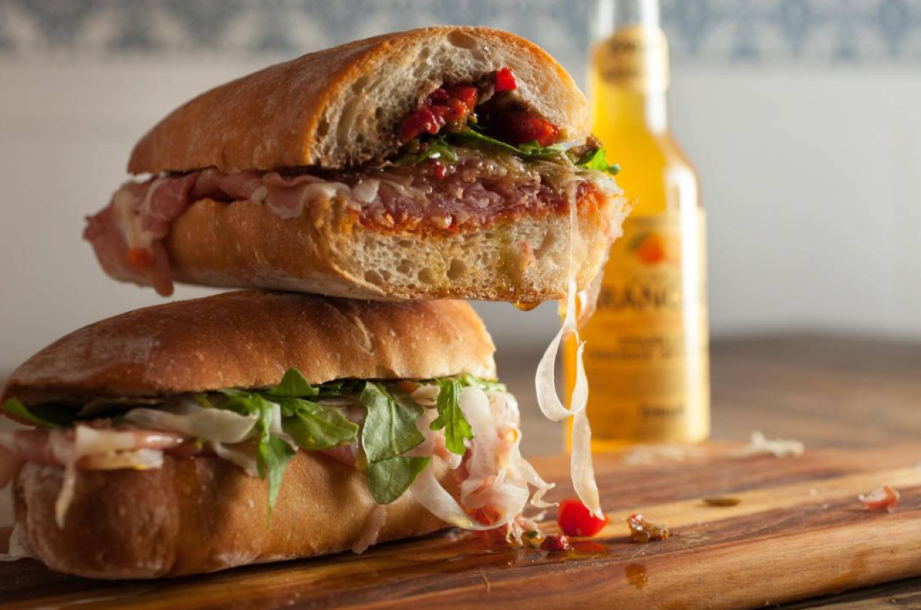 Two stacked ciabatta sandwiches layered with cured meat, greens, and peppers, reminiscent of restaurants across the USA known for next-level hoagies, with a bottle of orange soda in the background.