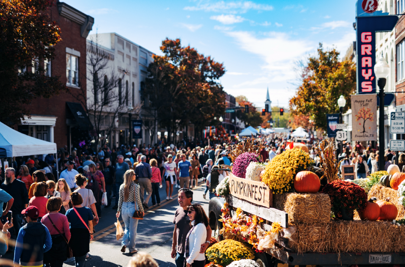 A crowded street festival with a sign that reads “PUMPKINFEST” on the back of a truck decorated with hay bales, pumpkins, and fall flowers.